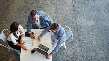 Image of three work professionals sitting at a table with a papers and a computer.