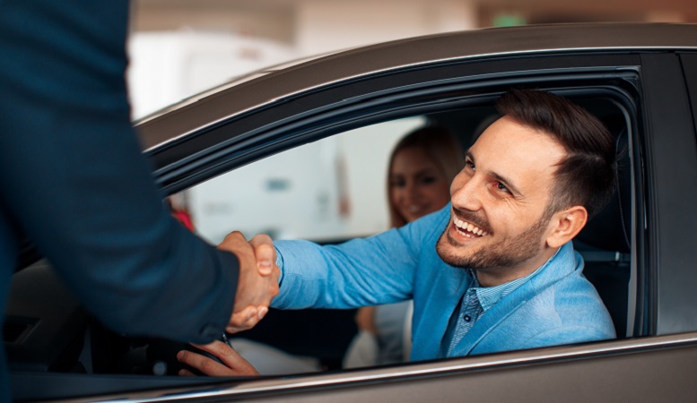 Two individuals shaking hands. One individual is in a car leaning out of the window and is smiling. In the background is a passage, also smiling while looking towards the individual outside of the car.